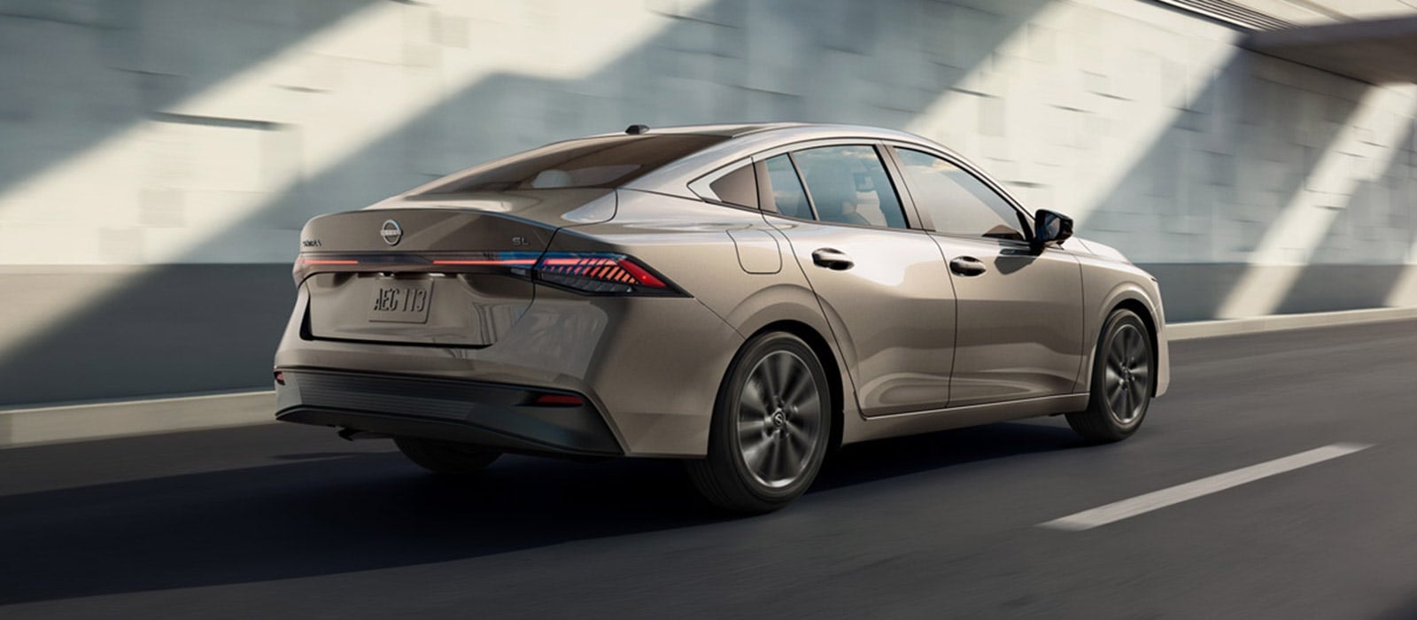 A tan Nissan Sentra driving down the road against a concrete wall.