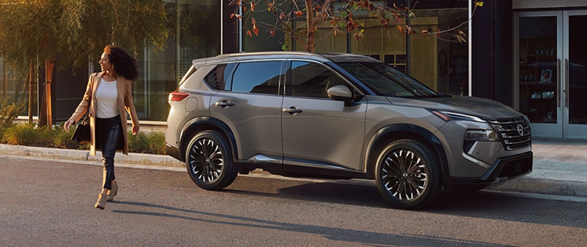A gray Nissan Rogue parked by a curb with a woman walking away from it.