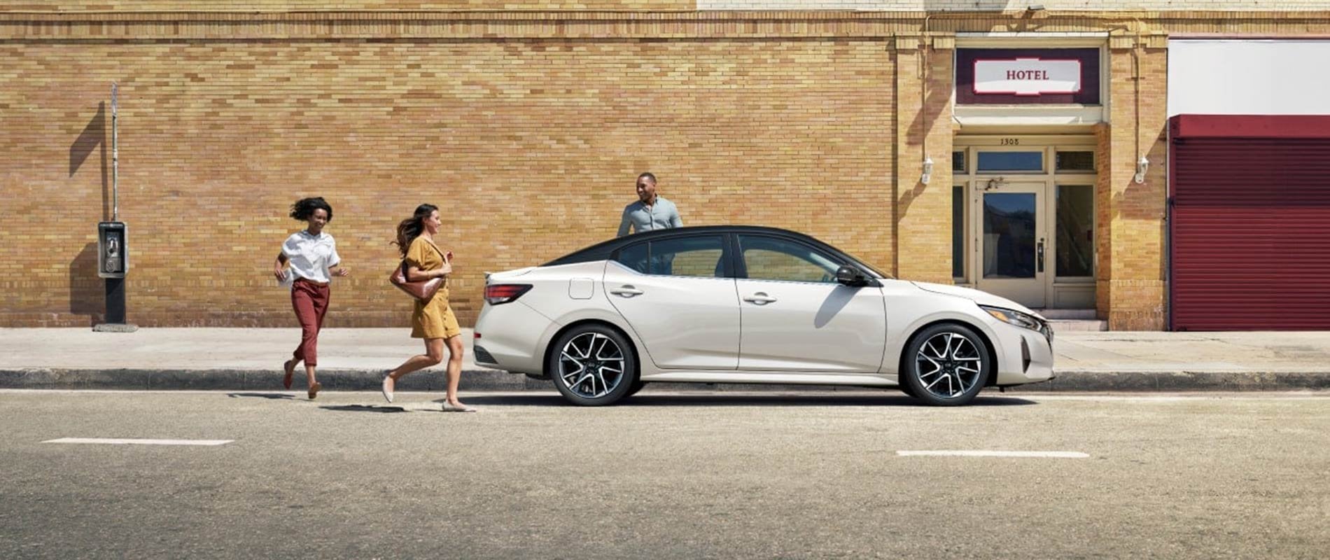 A profile image of a white Nissan Sentra parked by the curb of a hotel, as a trio of friends prepare to get inside it.